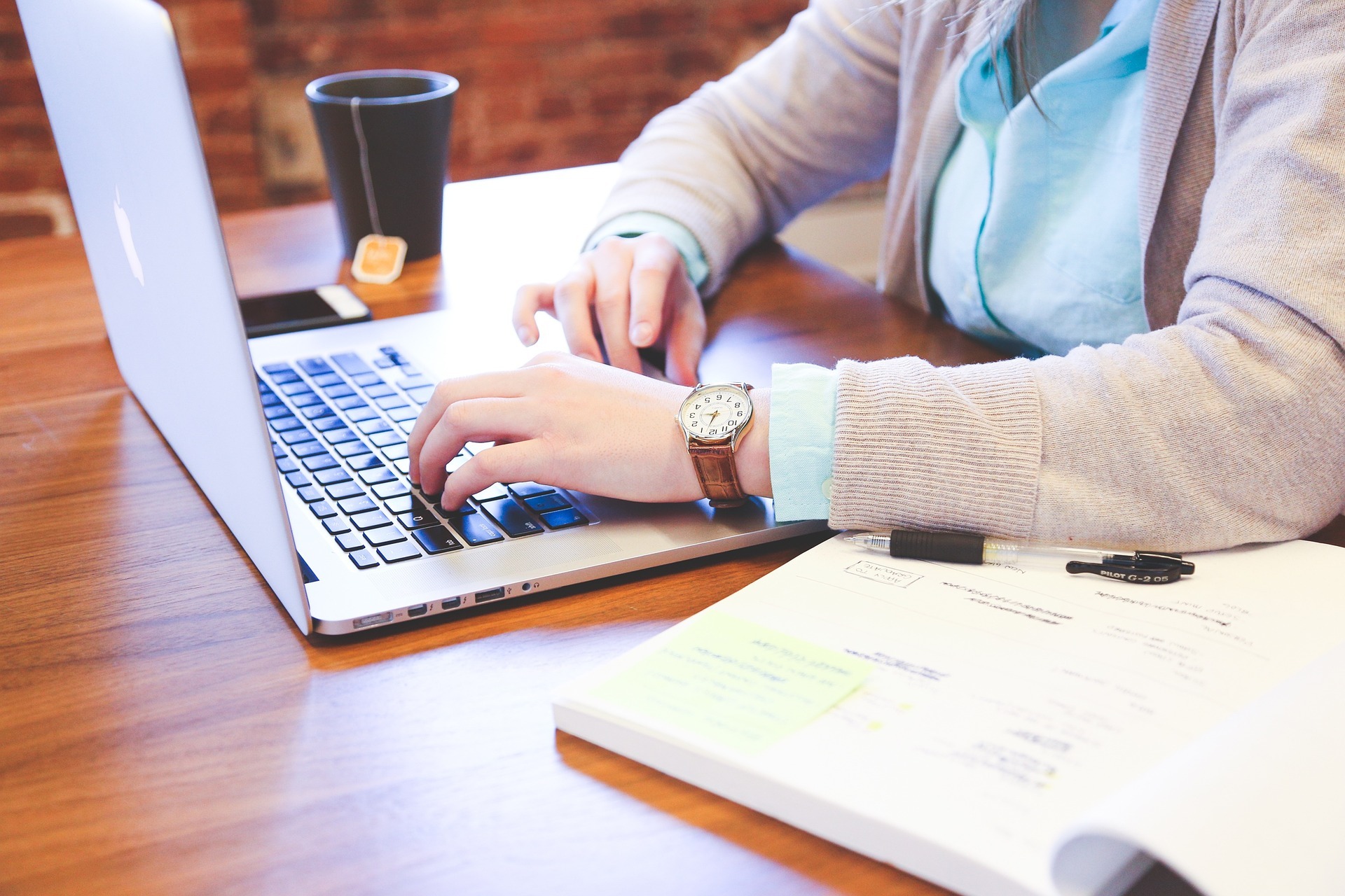 A woman wearing a watch and a pink sweater uses a Mac laptop on a wooden desk