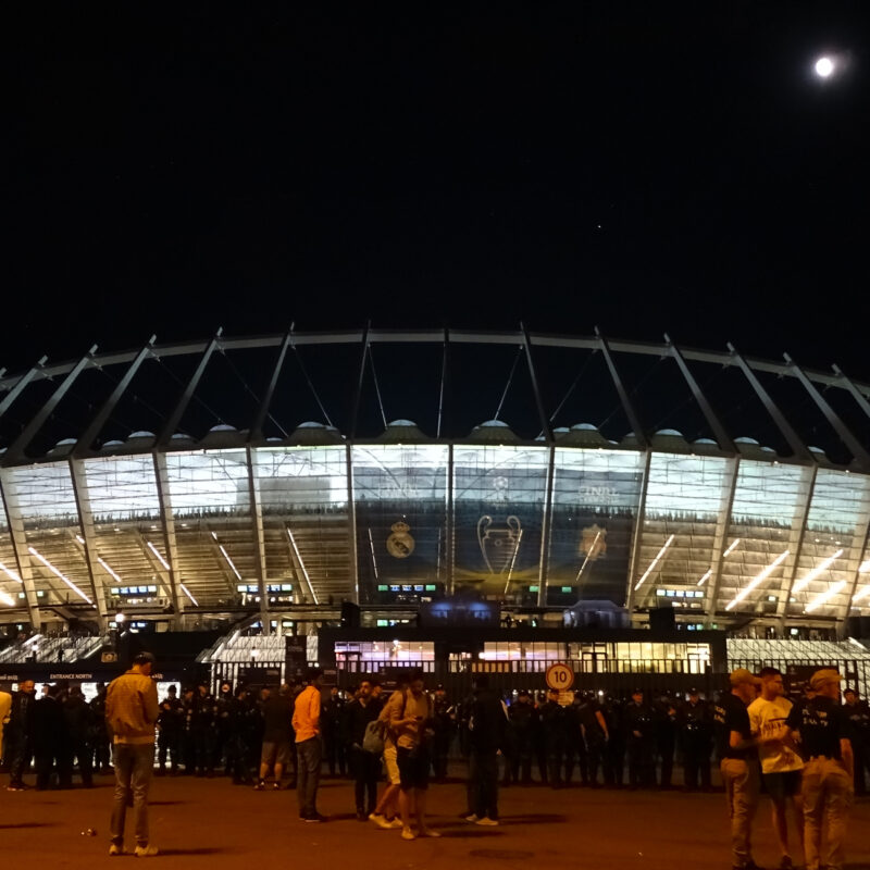 Wide night view of the Olimpiyskiy Stadium in Kyiv, Ukraine, with a small crowd gathered outside.